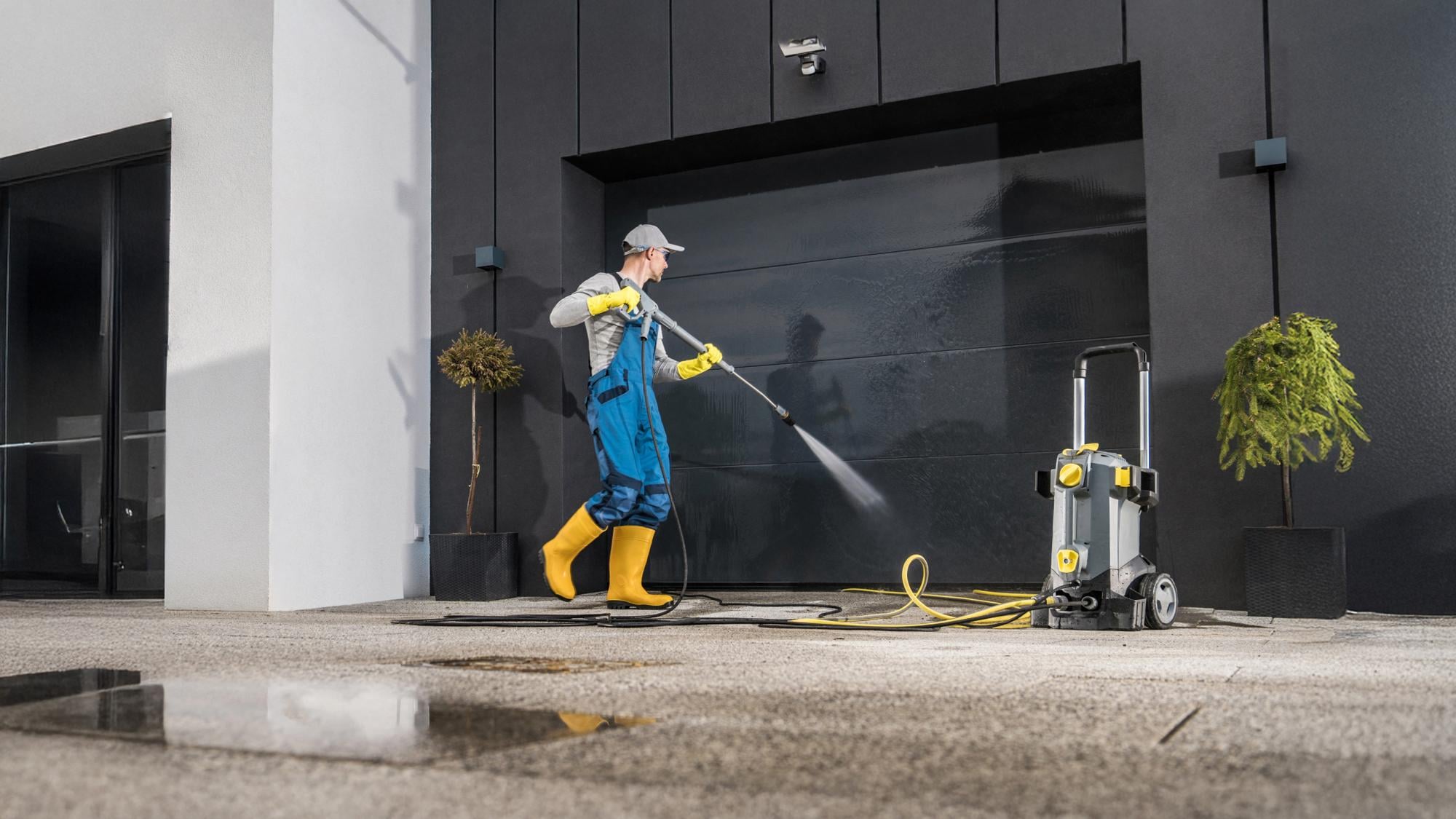 A person in blue overalls and yellow rubber boots is using a pressure washer to clean a dark garage door in a modern setting, with a potted plant nearby and wet pavement.